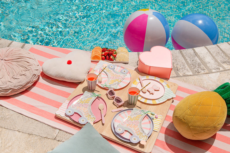 A pink striped towel lies by a pool in the sun, and rollerskate retro plates and tableware is placed on top beside several cushions including a large pineapple.