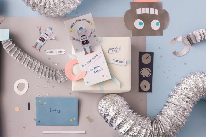 A flat lay featuring Father's Day crafts and gifts with a robot theme, including a "Happy Father's Day" card, a wrapped gift with a "I Love You A Bot" tag, and various metallic and paper robot parts.