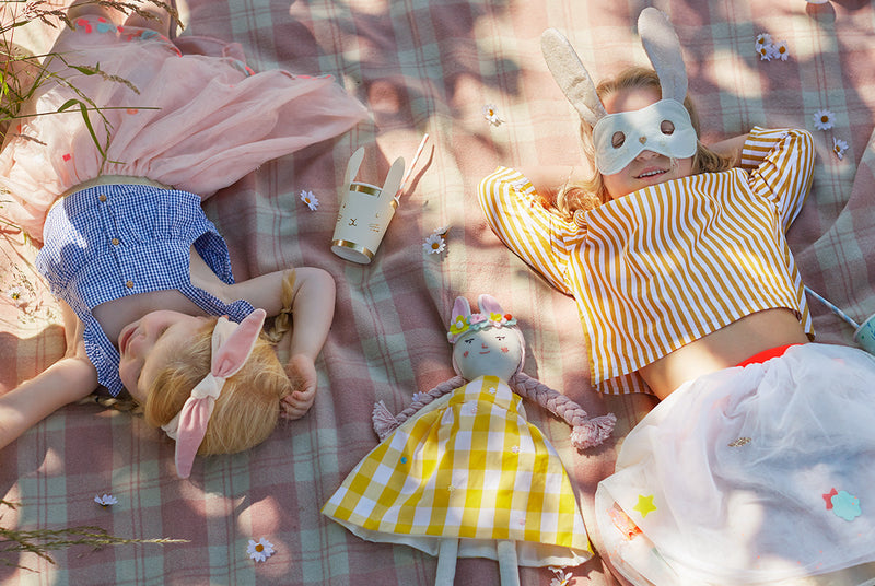Two children in Easter outfits lie on a pink plaid blanket, one with bunny ear headband and pink dress, the other wearing a white bunny mask amid Easter toys.