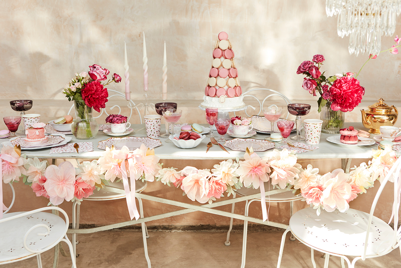 Pastel wedding dessert table with macarons tower, pink flowers, teacups, candles, and a floral garland.