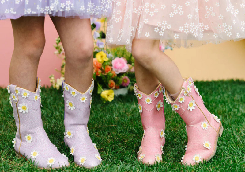 Girls wearing lavender and pink daisy embroidered boots stand on a grassy lawn with colorful flowers in the background.