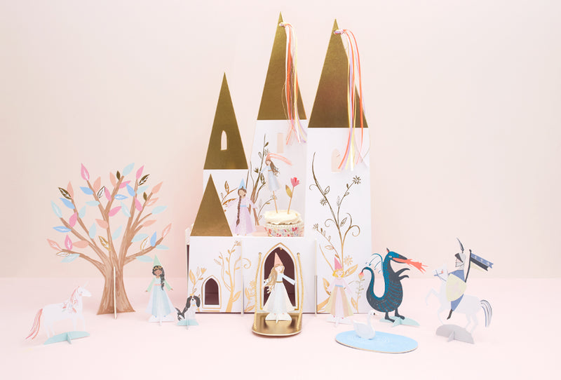 Pastel paper princess castle with gold-topped towers, surrounded by fairy figures, a blue dragon, and decorative trees.