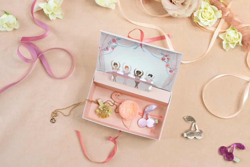 A dusty pink jewelry box with a line of ballerina characters along the top filled with accessories against a backdrop of light pink and ribbons.