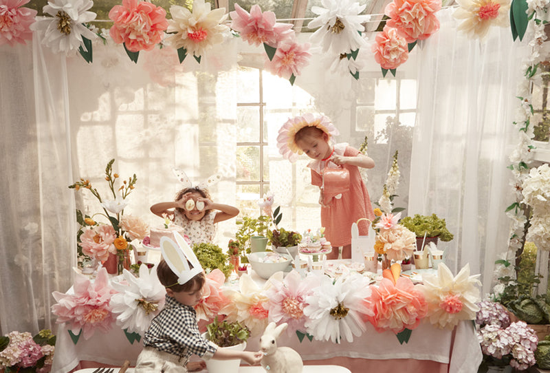 Three kids at a pastel Easter party table, surrounded by enormous paper flowers and greenery.