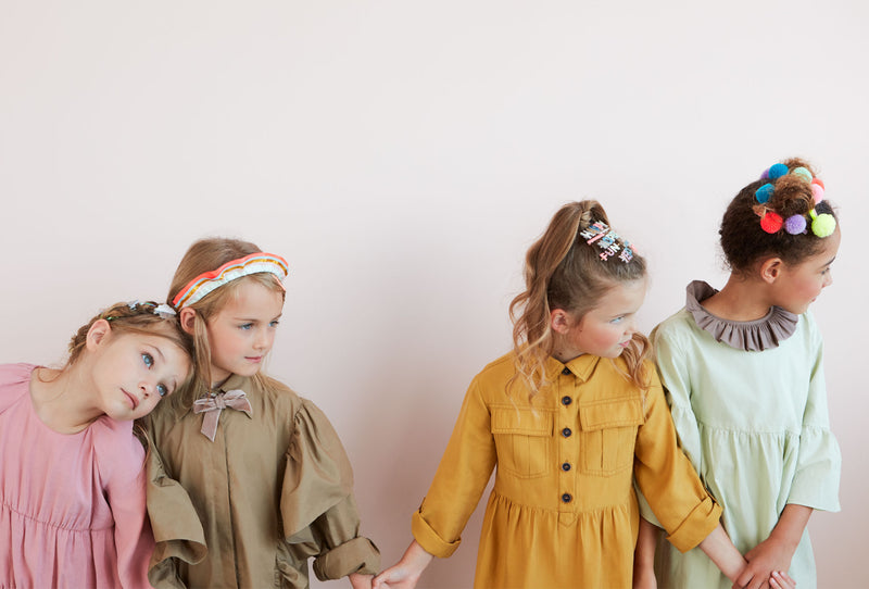 Four girls in colorful dresses pose against a pale pink wall, ready to craft friendship bracelets.