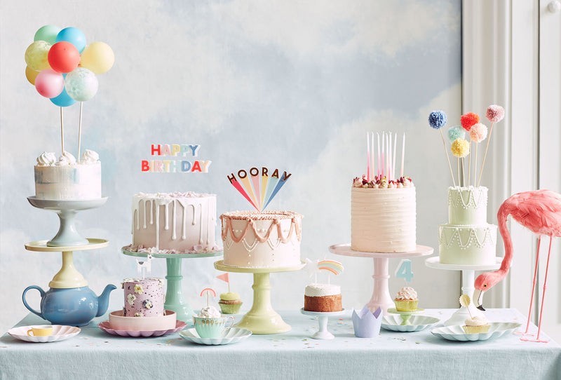 A line of large cakes covered in frosting placed on cake stands display a range of colorful candles and cake toppers.