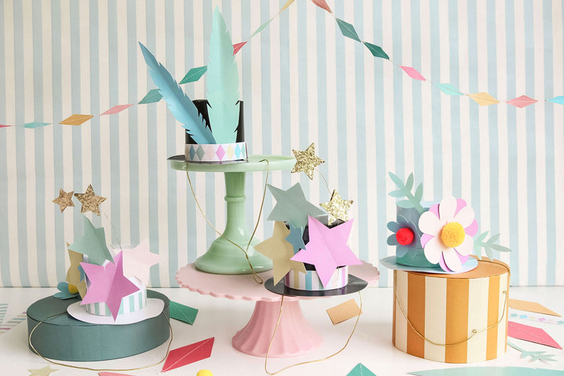 Various circus hats made from colored card and in different designs such as stars and flowers are displayed against a striped blue background and below a pastel diamond-shaped garland.