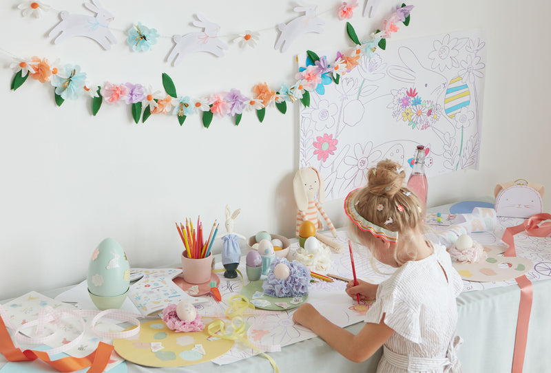 A young girl draws at a table covered with Easter eggs and stickers, with a Easter coloring poster above and floral garlands.