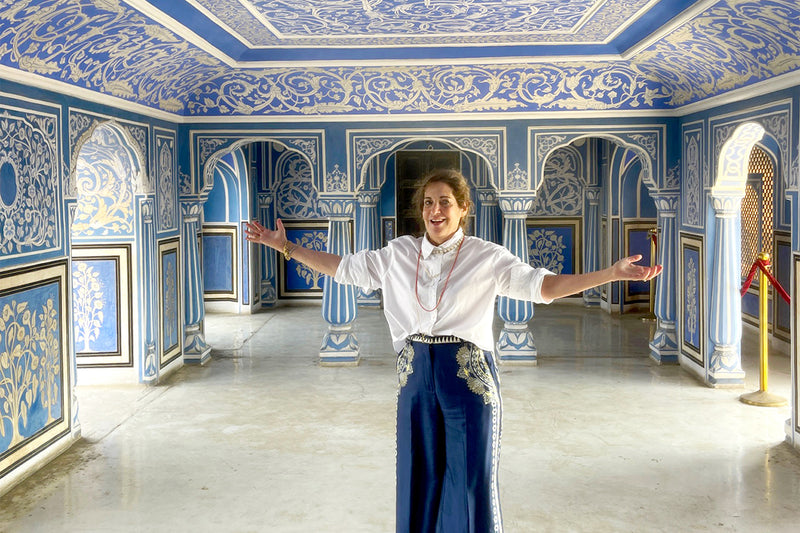 Tourist in a white blouse and blue embroidered pants stands with arms outstretched inside a blue and white ornate Jaipur palace interior.