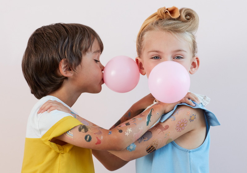 Two kids pose with pink balloons, one kissing the other's balloon while colorful temporary tattoos show on their arms.