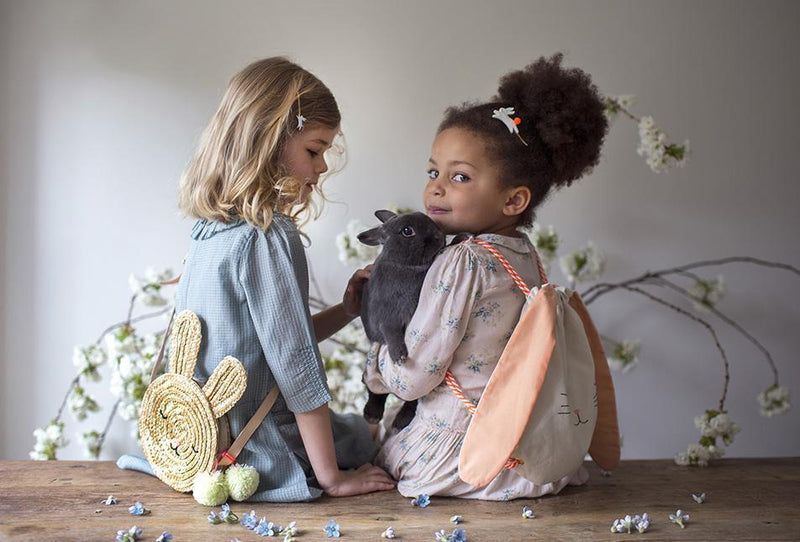 Two girls in pastel dresses sit on a wooden surface, each with a playful animal backpack in a party styled scene.