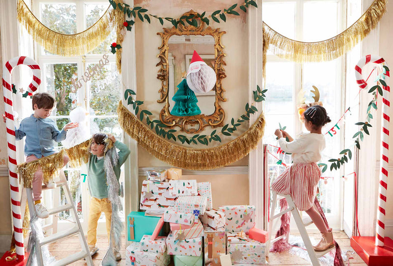 Children decorating a sunlit doorway with candy cane arches, gold fringe garlands and festive paper ornaments around a gilded mirror