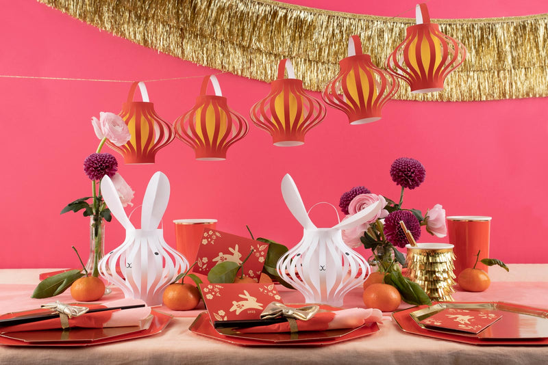 Party supplies inspired by the Lunar New Year are laid on a table, including red plates, bunny lanterns, and red and gold napkins. A gold tinsel banner and red lantern garland hang from the ceiling.