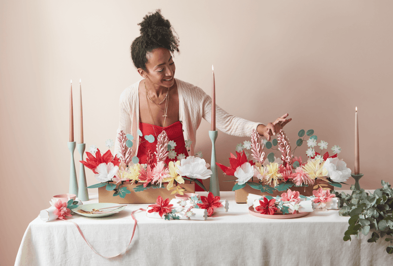 Woman in a light cardigan shapes a festive pastel floral centerpiece with tall pink candles at a decorated table