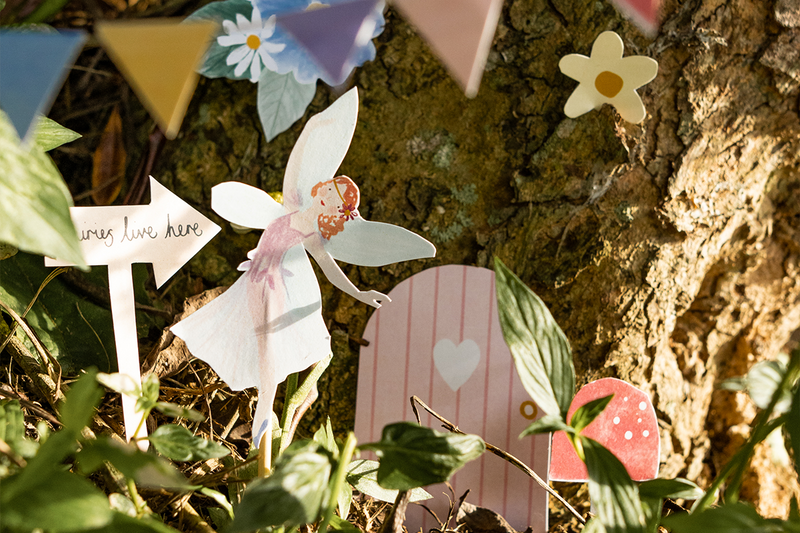 A paper fairy cut-out stands in front of a tree trunk with a little door and mushroom, creating a fairy garden out of printable designs.