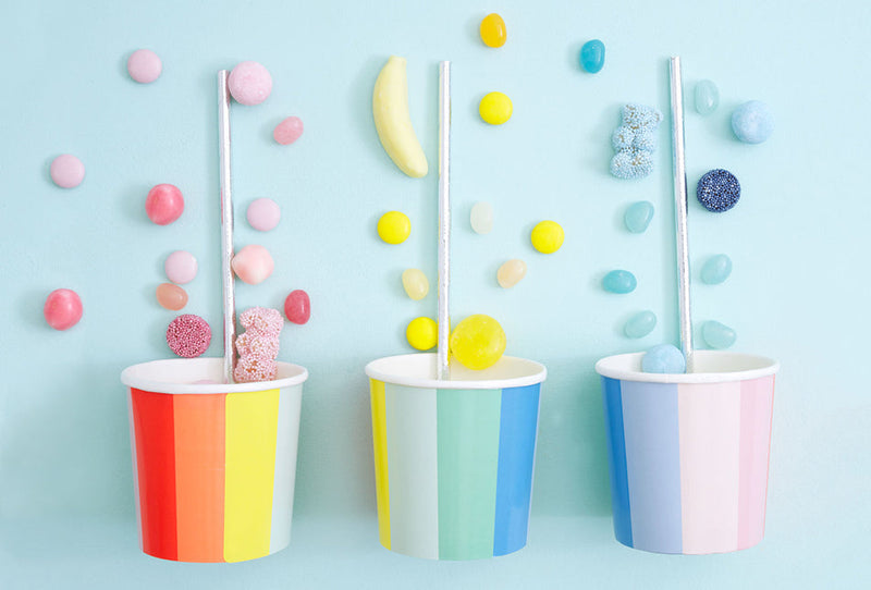 Three rainbow striped cups with silver straws on a pastel blue background, colorful candies spilling around them.