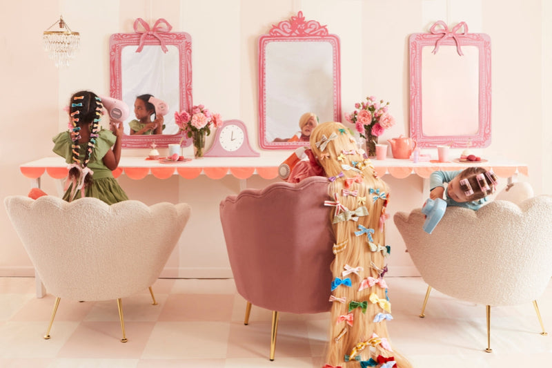 Girls in a pastel pink hair salon with large mirrors, styling hair with curlers and ribbons.