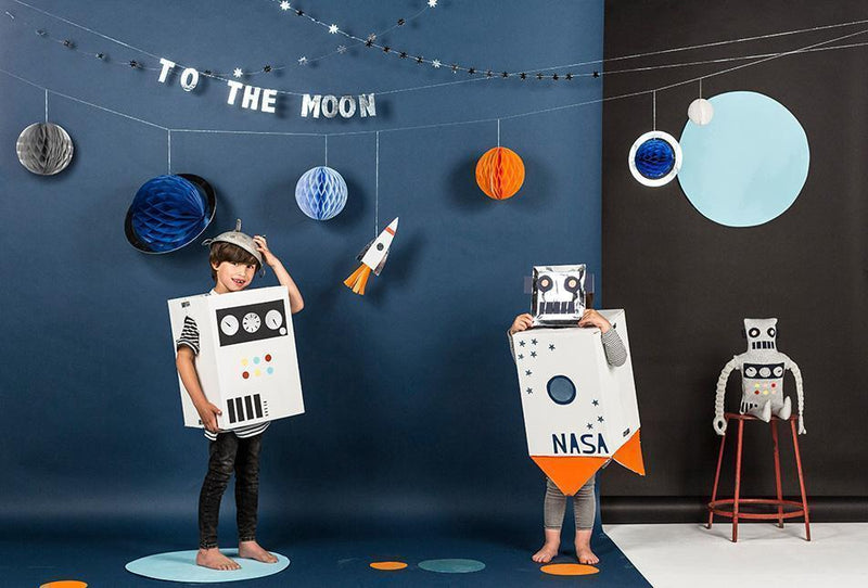 Two children in cardboard rocket costumes pose beside a space themed backdrop with planets and a robot prop.