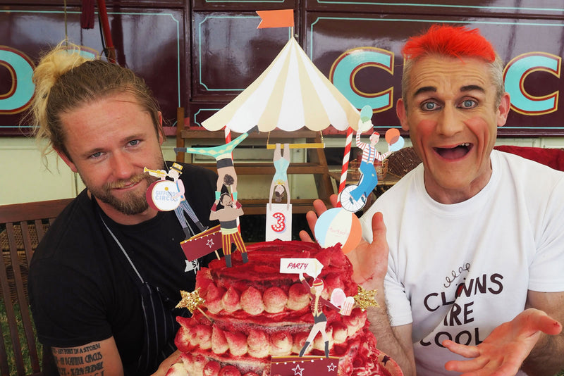 Two members of Gifford's Circus show off a red cake topped with circus characters and a big top, including strong men, acrobats, and clowns.