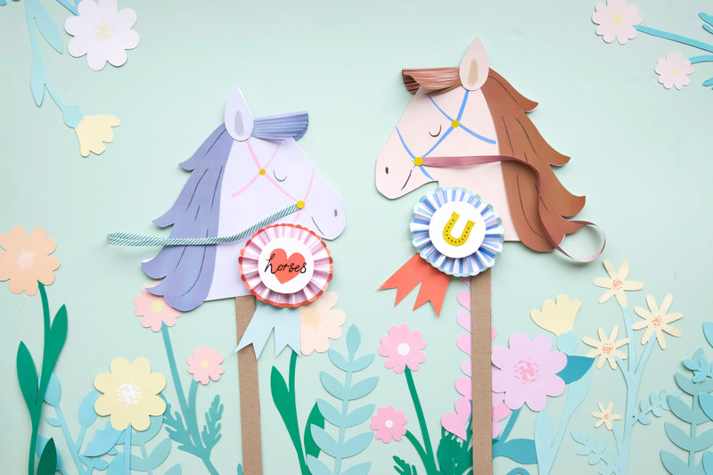 Two handmade paper stick horses with ribbons and rosettes against a pastel floral background