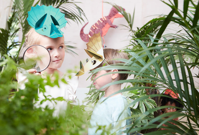 2 young children wear dinosaur hats and are surrounded by green leaves and plants whilst a third child hides in the background.