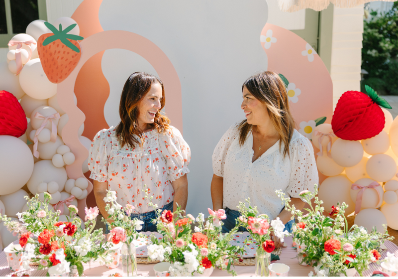 Jacquelyn Kazas of Beijos Events standing behind a table adorned with a variety of floral decorations and desserts, looking at each other and smiling. In the background, there's a strawberry-themed wall with balloons and strawberry cutouts.