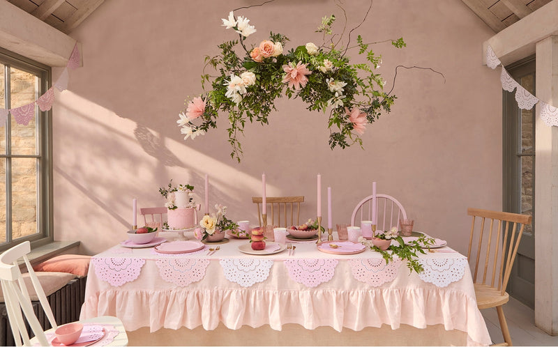 A beautifully decorated table in a room with pink walls, featuring pink tablecloths, plates, candles, and a hanging floral arrangement with roses and other blossoms.