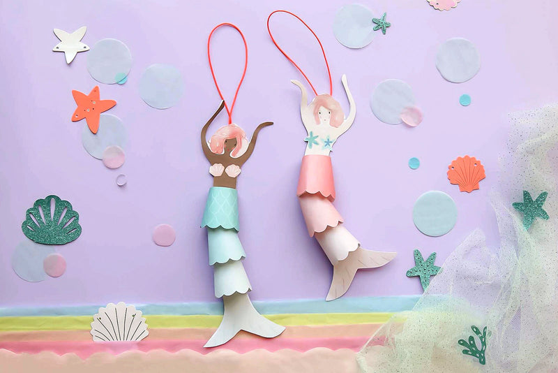 Two DIY mermaid ornaments made out of blue and pink card, left to right respectively, appear against a purple background featuring seashells and bubbles. 