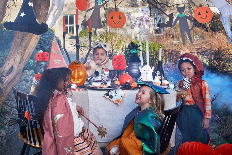 4 children wearing Halloween costumes, including a dinosaur costume and a witch's outfit, sit around a table decorated with Halloween party supplies such as honeycomb pumpkins and spiders, and a garland with Halloween motifs hangs above.