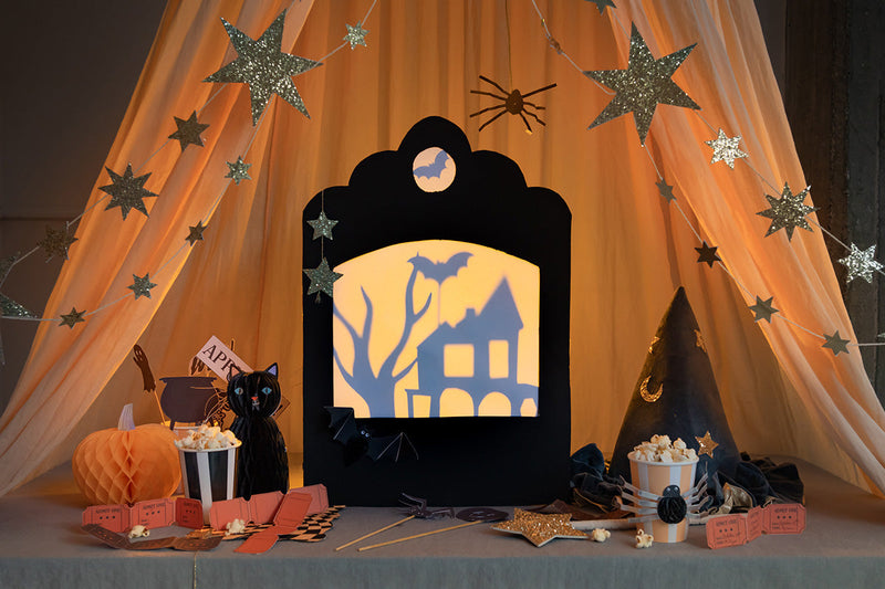A small cardboard shadow theater with Halloween-inspired shadows sits under a star garland and orange tulle, and is surrounded by honeycomb pumpkins and cat, a witch's hat, and striped cups with popcorn.