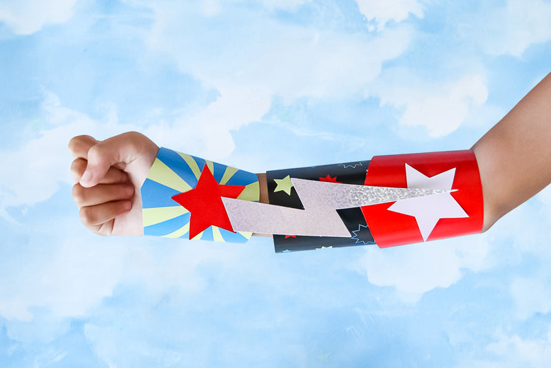 In front of a background of the sky, an arm displays a DIY superhero cuff made with colored card and finished with stars and a lightning bolt.