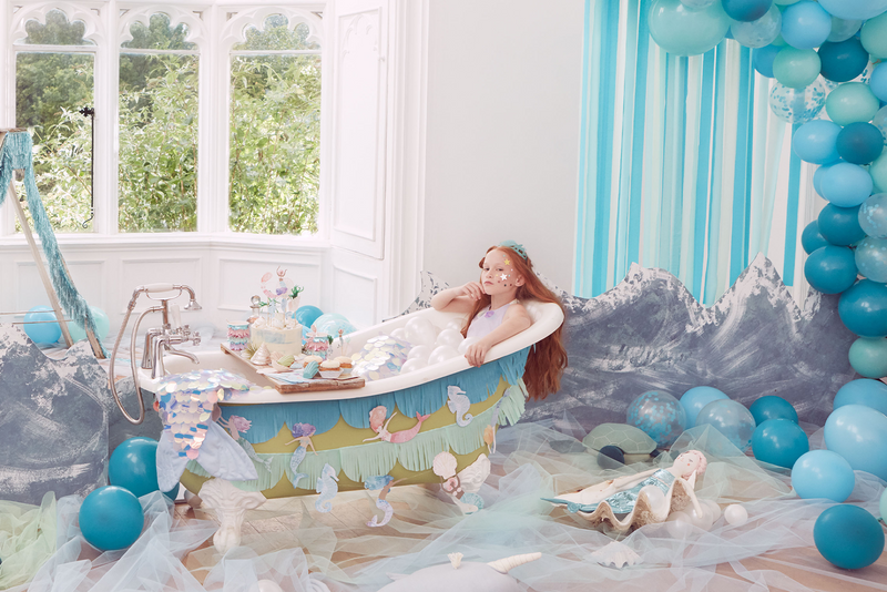 A young girl with red hair sits wearing a mermaid tail and top in a bath with mermaid crepe banners on the outside. Blue paper streamers and a statement blue balloon arch feature in the background.