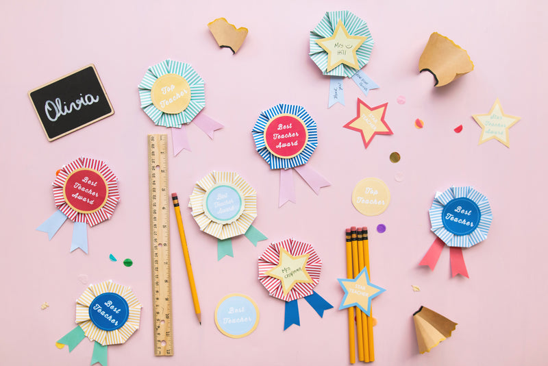 Paper rosettes in a mix of yellow, blue, and red that read 'Best Teacher' are featured against a pink background.