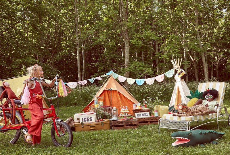In a forested area, a young child sits on a red bike with a monkey toy on the back and another child sits on a chair with a leopard and crocodile toy.