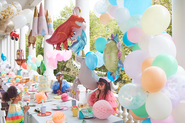 Three children sit at a long table decorated for a dinosaur-themed party, surrounded by colorful balloons and dinosaur decorations on a porch.
