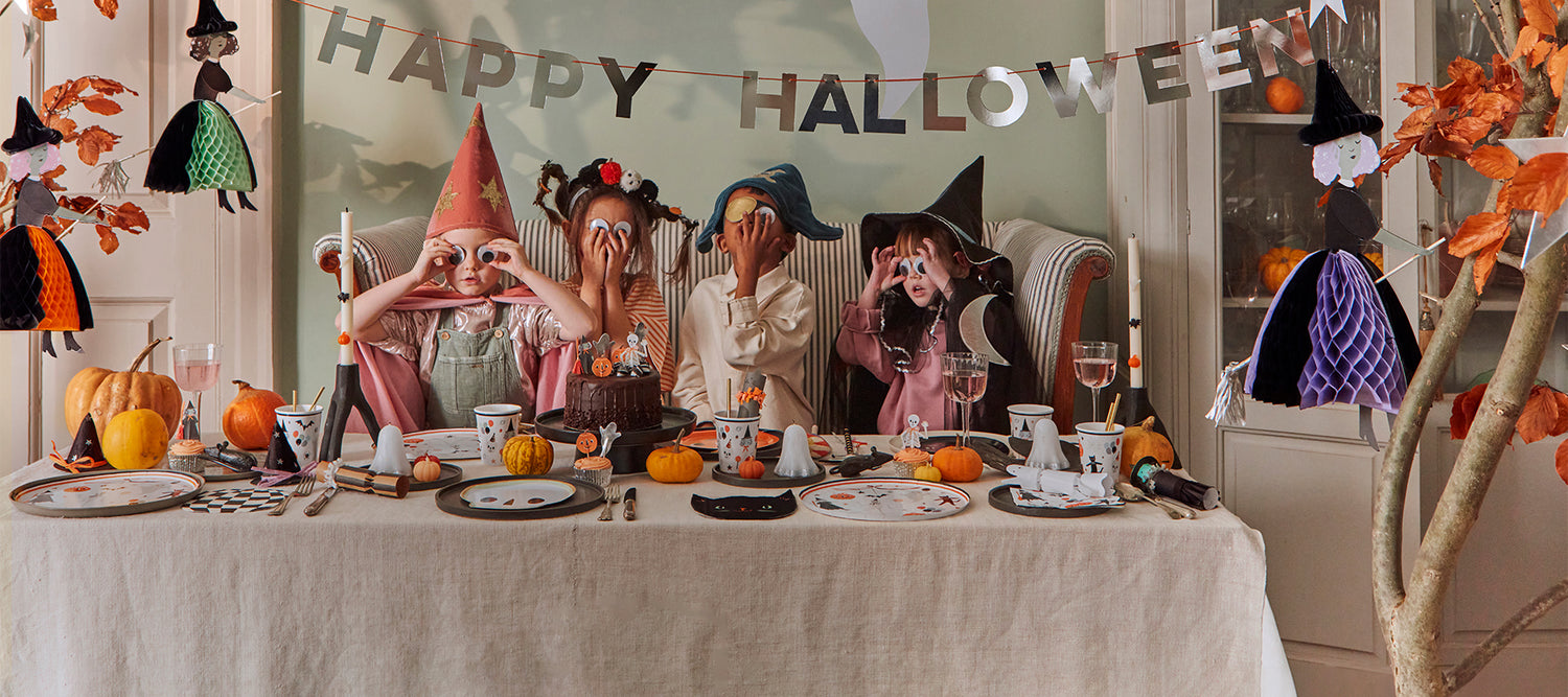 Kids wearing Meri Meri Halloween costumes and googly eyes, sit next to a Halloween party table that is decorated with fun tableware and pumpkins. 