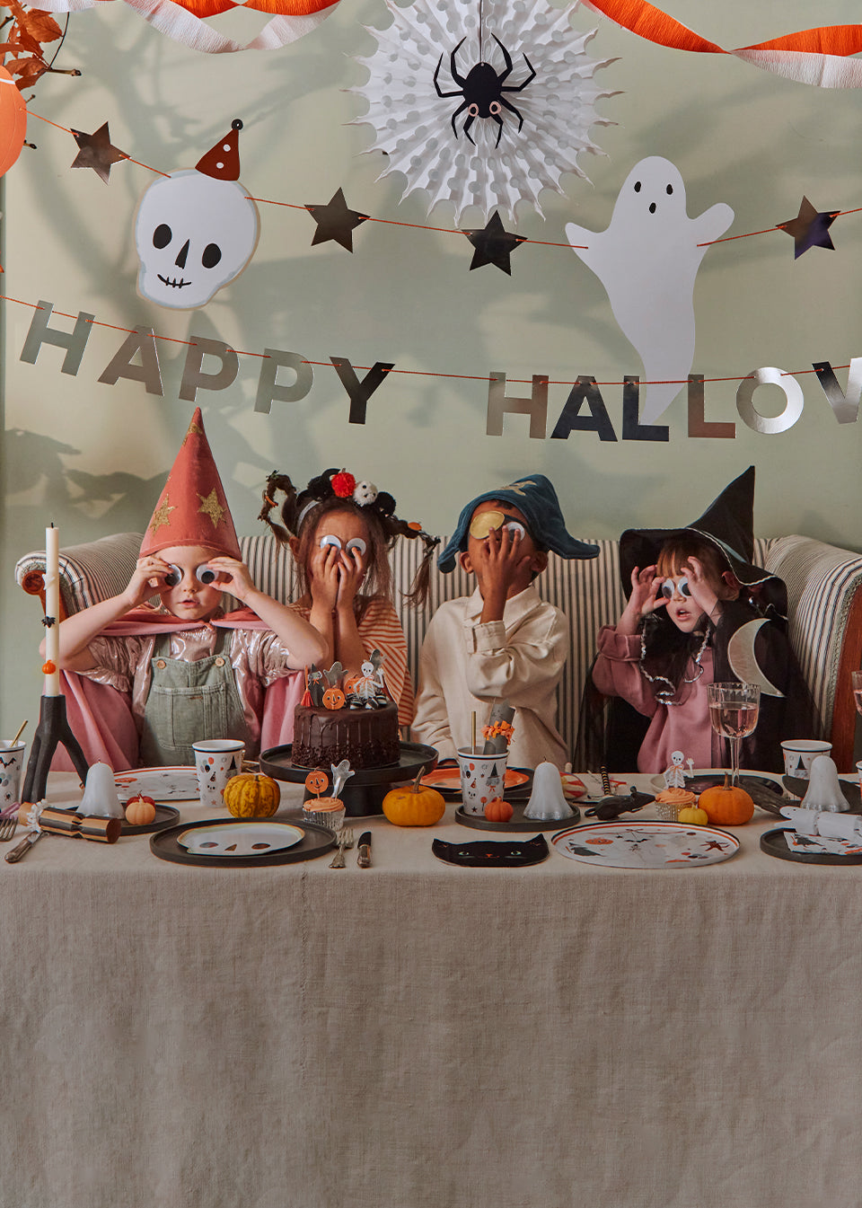 Kids wearing Meri Meri Halloween costumes and googly eyes, sit next to a Halloween party table that is decorated with fun tableware and pumpkins. Mobile