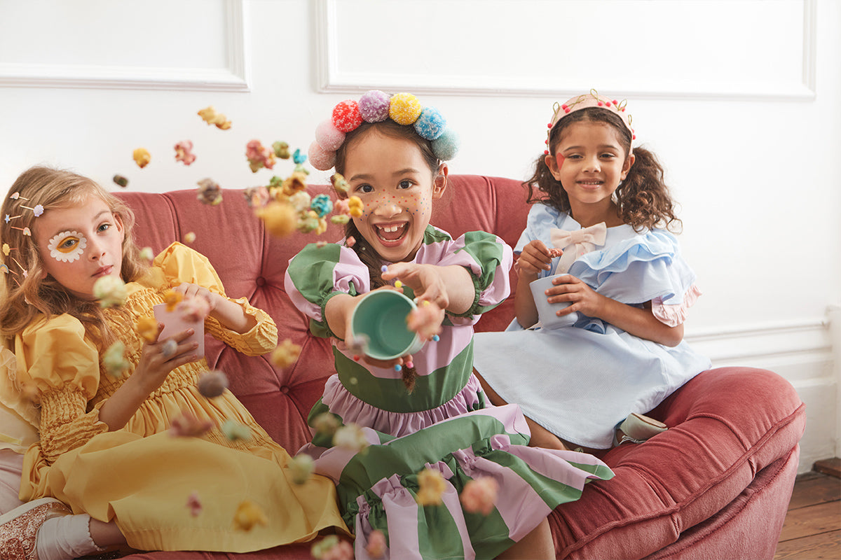 3 young girls sit on a sofa in colorful outfits with hair accessories whilst the middle throws a cup of colored popcorn at the camera.