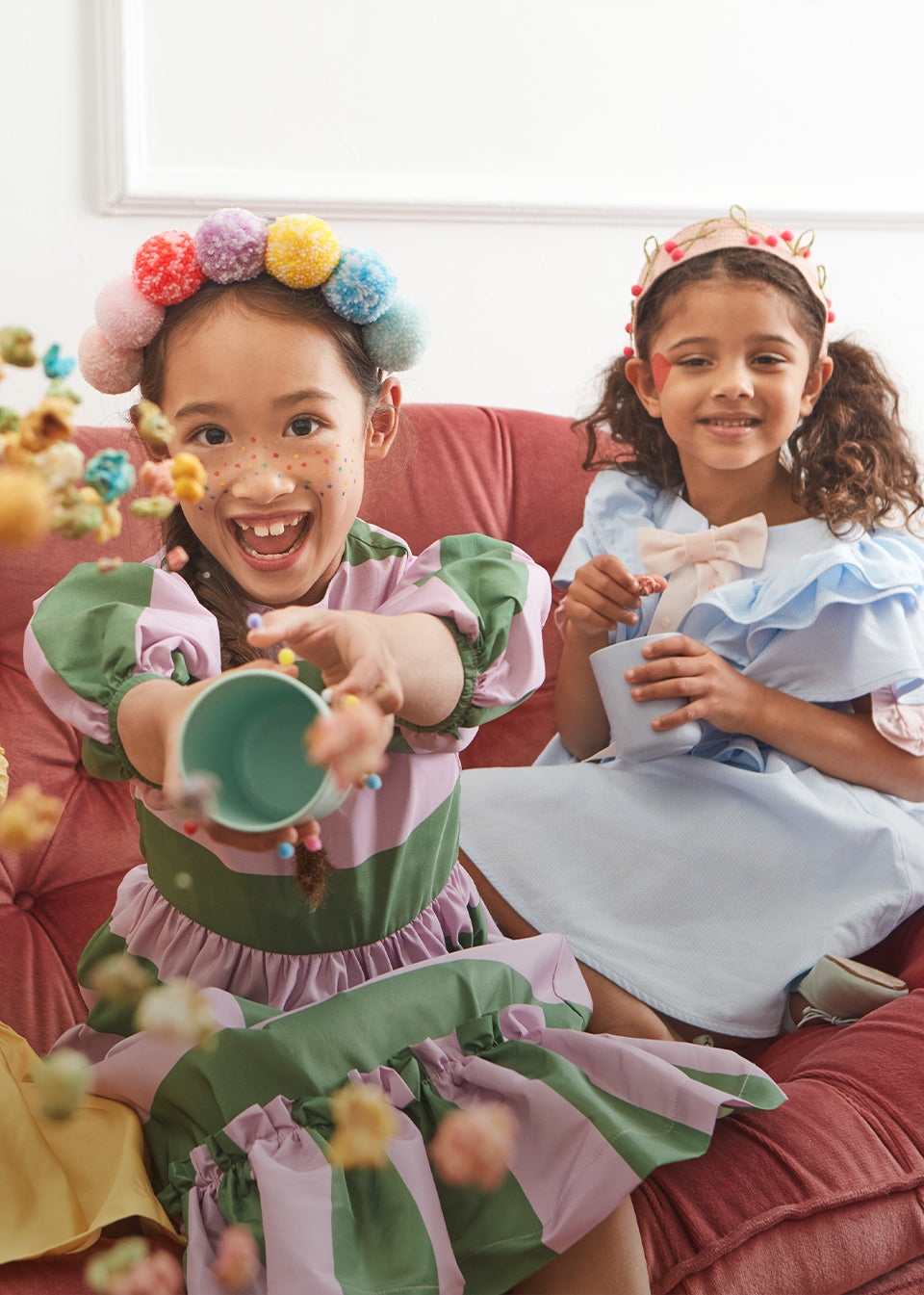2 young girls sit on a sofa in colorful outfits with hair accessories whilst the girl on the right throws a cup of colored popcorn at the camera.