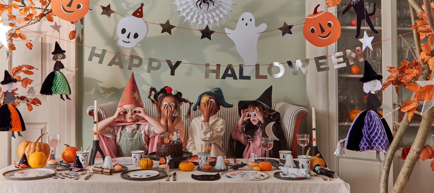 Four kids having a fun Halloween party dressed in Meri Meri costumes sat around a table adorned with Meri Meri party supplies. 