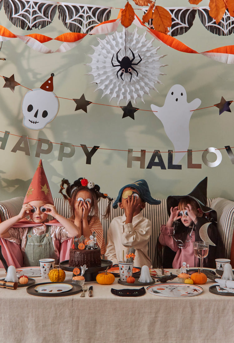 mobile friendly image of Four kids having a fun Halloween party dressed in Meri Meri costumes sat around a table adorned with Meri Meri party supplies. 