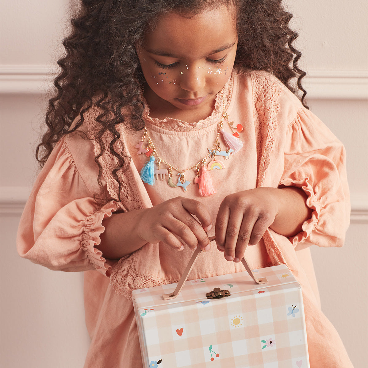The pink gingham mini suitcase opens to reveal numbered trays filled with enamel charms, tassels and a gold tone necklace.