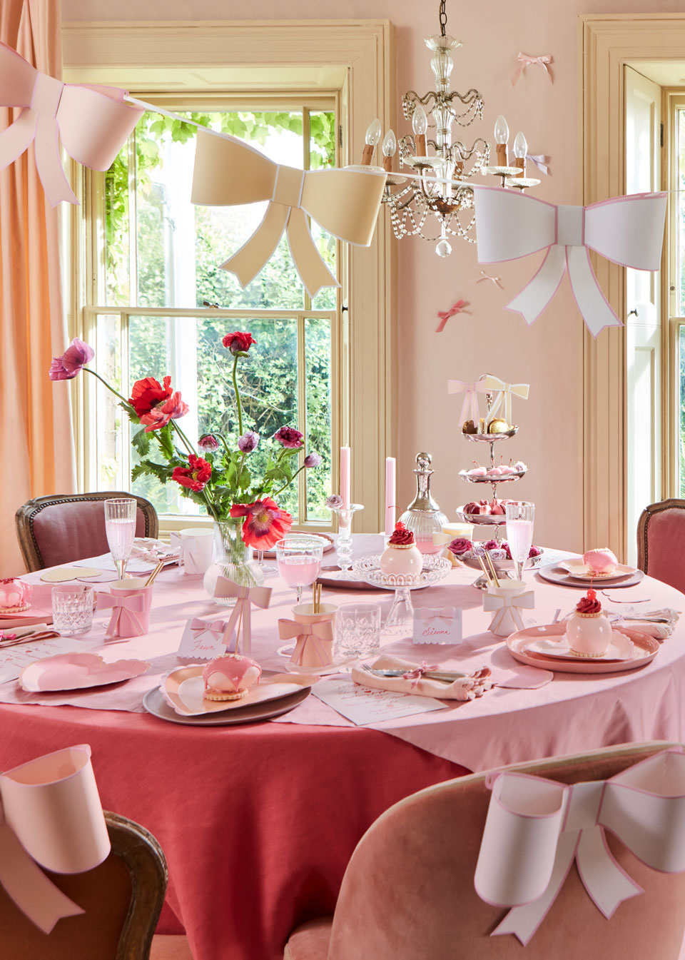 In a pink-themed dining room, a string of bows in pastel shades of pink, yellow, and blue hangs in the foreground whilst a table surrounded by dusty pink chairs is bedecked in pink heart plates and tableware accessorized with bows.